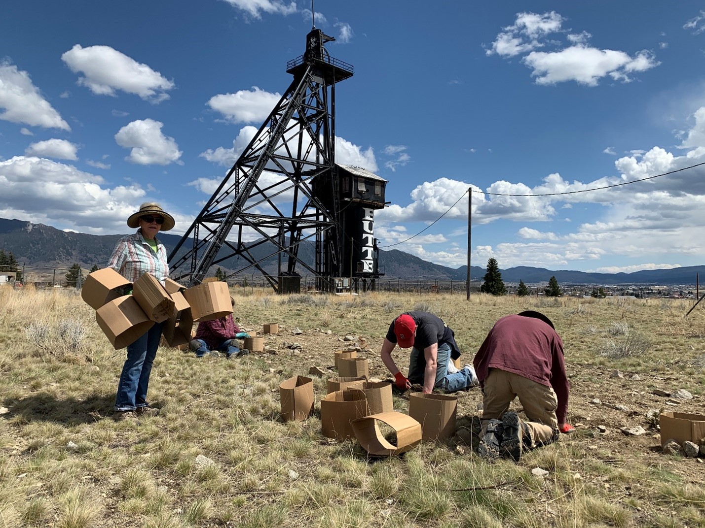 A group of people engaged in a cleanup activity outdoors near a historic mining structure, with mountainous scenery and a partly cloudy sky in the background.