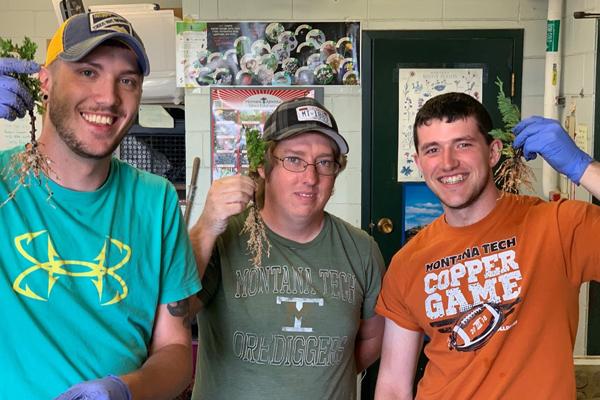 Three individuals are smiling and holding up plants with roots, showcasing their work in agriculture or botany.