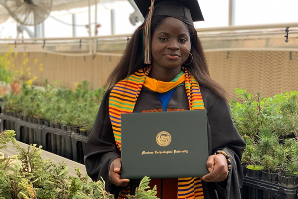 A graduate holding a diploma from American International University while surrounded by greenery in a greenhouse setting.