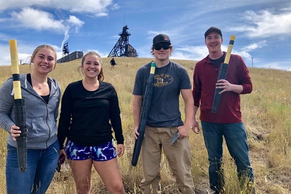 Four individuals stand in a field holding equipment, with a backdrop of tall grass and structures on a hill.
