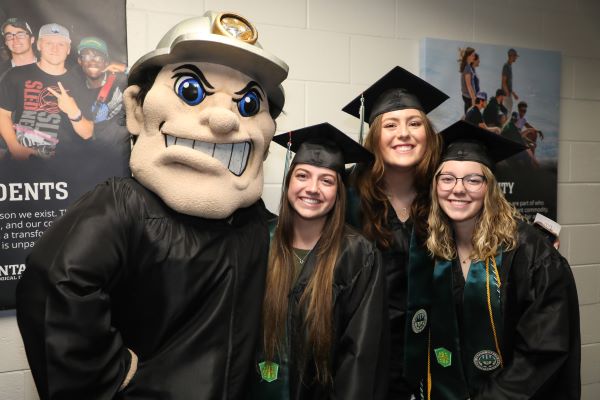 Three students pose with mascot Charlie Oredigger in their caps and gowns