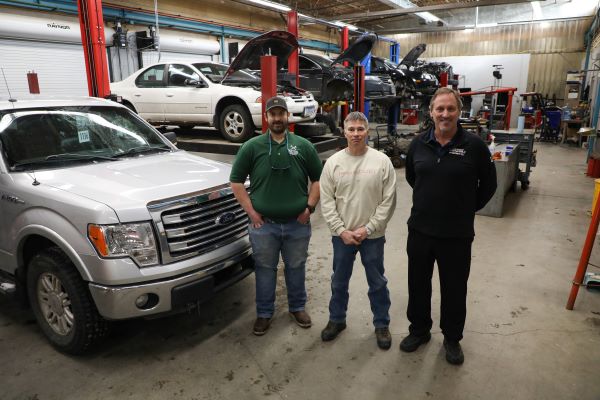 !Three men stand in front of vehicles in a workshop