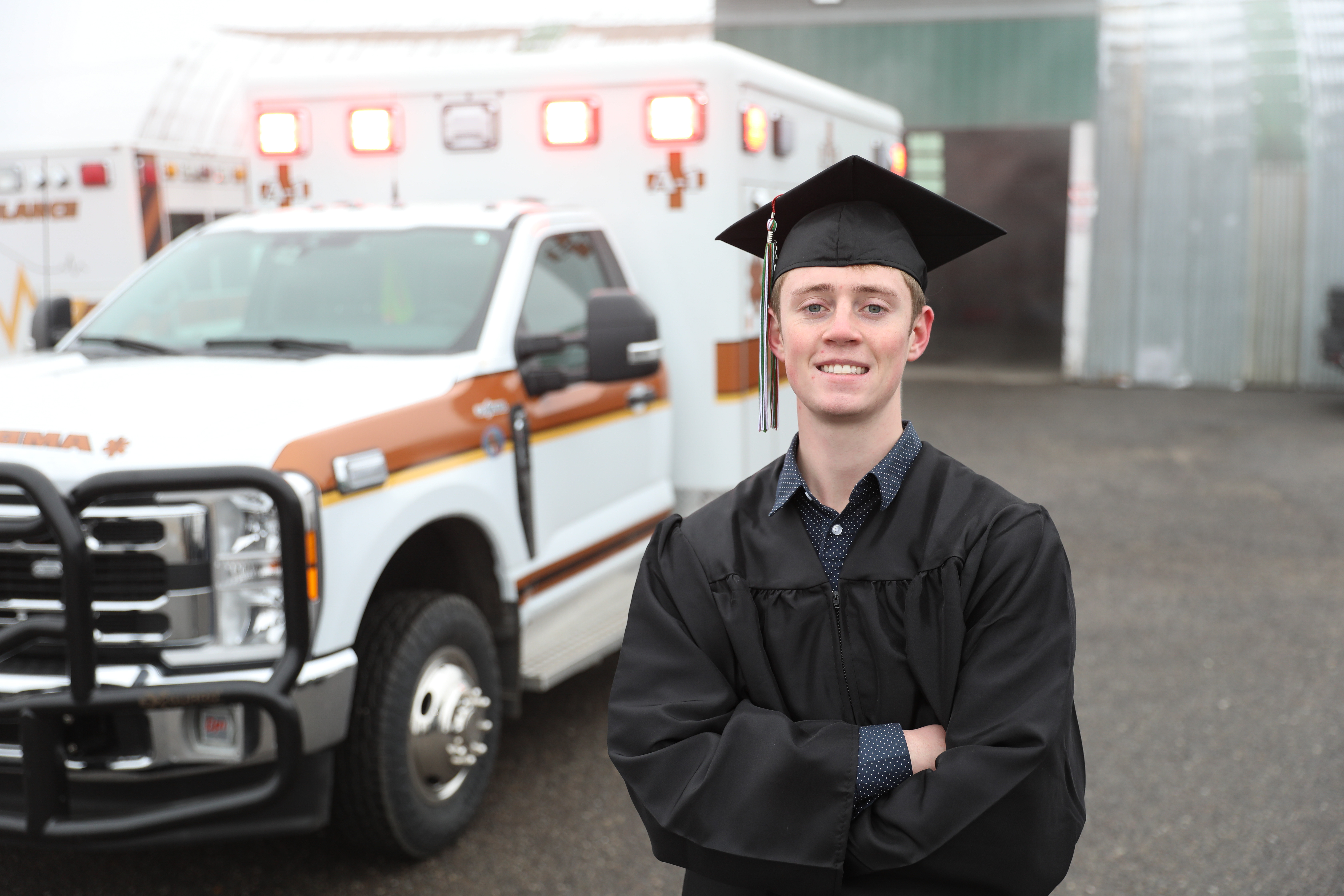 !Trey Whitlock in front of an ambulance in his cap and gown