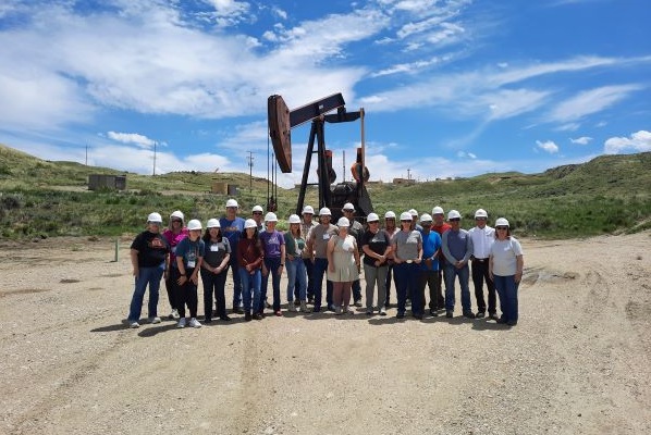 A group of teachers stand in hard hats in front of a pump jack