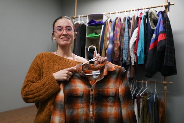 !Cheyenne Crooker holds up a plaid shirt in front of racks of clothing