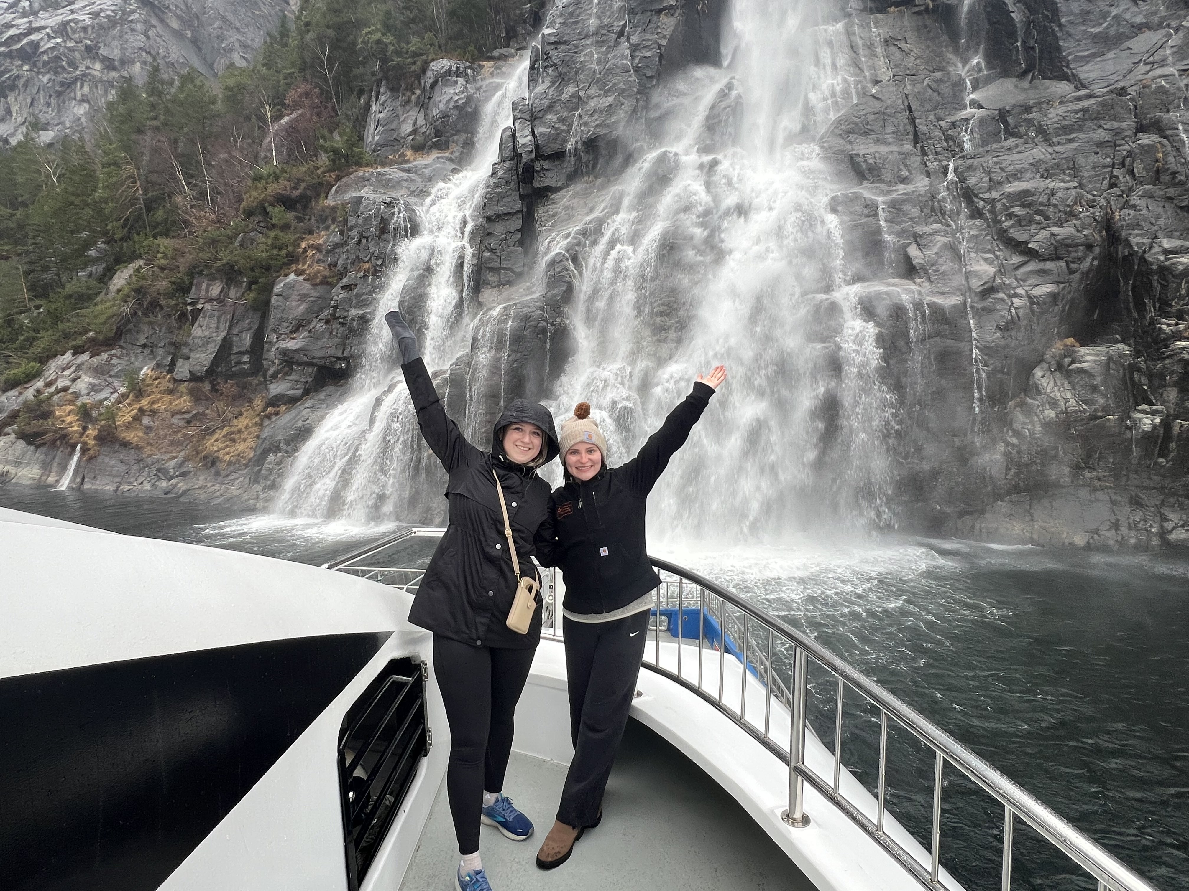 Two students pose in front of a waterfall on a cruise in a fjord